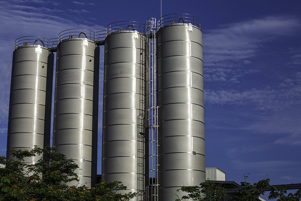 Large stainless steel industrial storage tanks at a chemical or food processing facility against a blue sky — stationary emission sources subject to EPA air permitting, environmental compliance, and regulatory consulting services by EDGE Engineering.