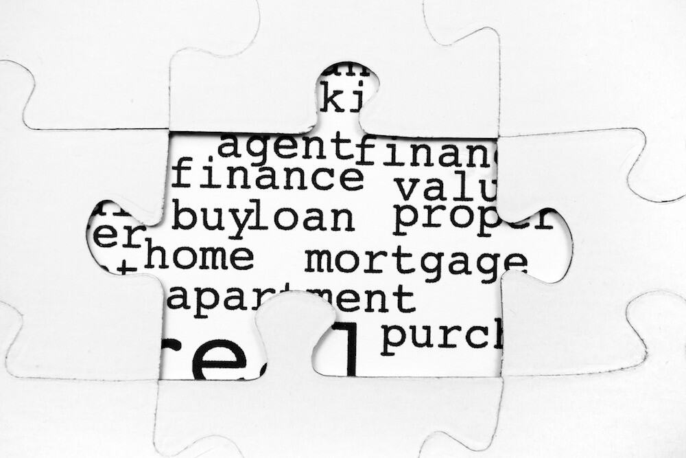 Conceptual black and white image of a missing jigsaw puzzle piece revealing real estate keywords like "mortgage," "home," and "finance" underneath.