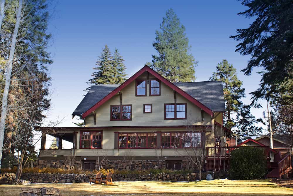 Large Craftsman-style home with sage green siding, red trim, and a stone foundation featuring a spacious lawn and tall evergreen trees.
