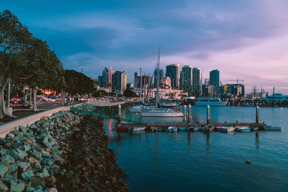 A panoramic view of the Downtown San Diego skyline across the water from the Downtown Harbor, featuring sailboats docked at a pier at sunset.