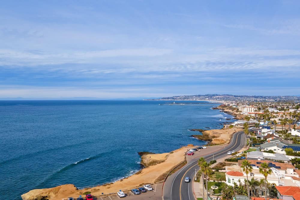 Aerial view of the Ocean Beach coastline in San Diego, featuring a winding coastal road, rocky cliffs, and the Pacific Ocean under a blue sky with light clouds.