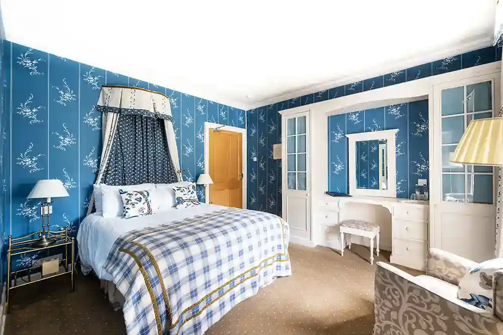Classic hotel bedroom with blue floral wallpaper, canopy double bed, white dressing table, and plaid bedspread.