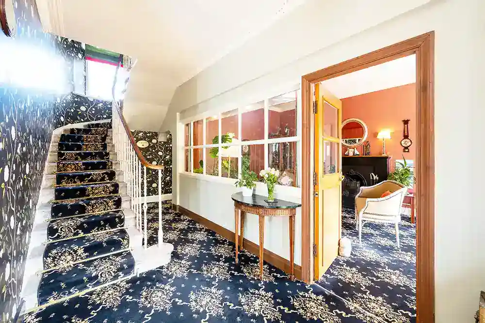 Grand hotel hallway with ornate navy patterned carpet, curved staircase, and floral console table.