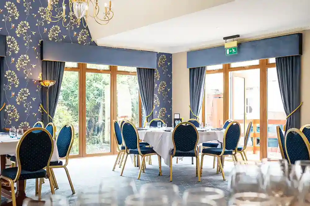 Bright hotel dining room with navy and gold chairs, floral wallpaper, chandelier, and garden-view windows.