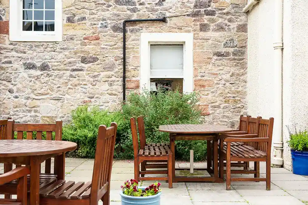 Rustic wooden patio tables and chairs against a stone wall with lush green garden shrubs.