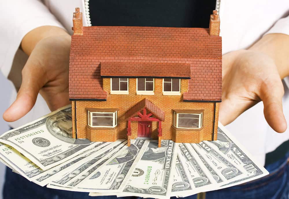 A person holding a model house sitting on top of a fan of US dollar bills, illustrating real estate investment or home purchase costs.