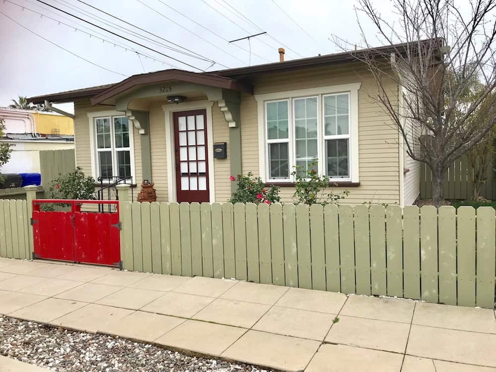 Exterior view of 3215 Myrtle in North Park featuring a tan bungalow with a red front door, a light green picket fence, and a vibrant red gate.