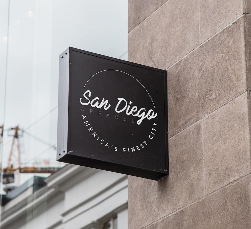 A square black outdoor sign mounted on a textured stone wall reads "San Diego Apparel, America's Finest City" in cursive and block lettering