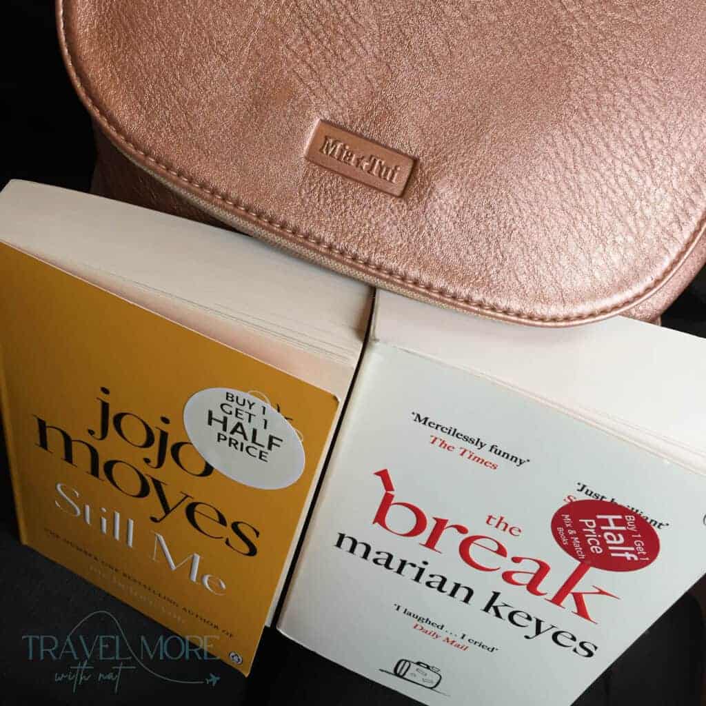 Two paperback books titled "jojo moyes Still Me" and "the break marian keyes" beside a metallic pink pouch. Books and small personal items like these are typically permitted under standard hand luggage rules.