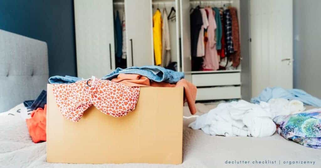 A box filled with clothes sits on a bed. Clothing is strewn across the bed. An open closet is in the background.