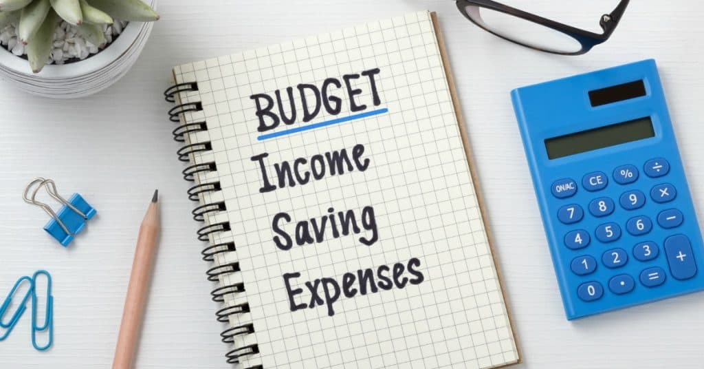Stylized desk with notebook, calculator, glasses, paperclips and small plant. Notebook says "BUDGET: Income, Savings, Expenses"