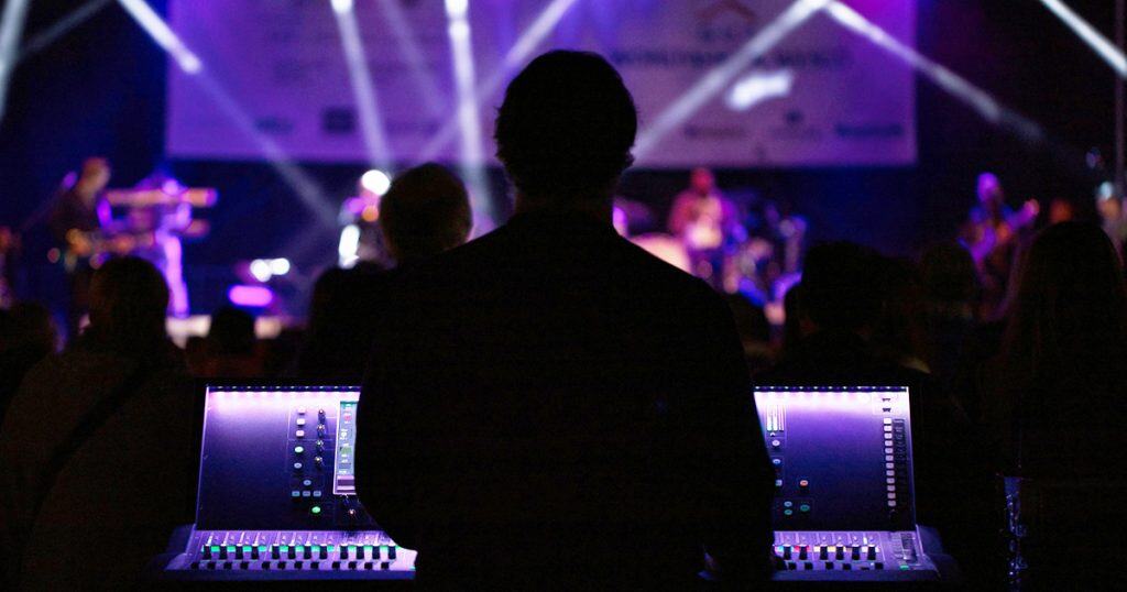 Live concert sound engineer working with digital audio mixing console at music event, vibrant stage lighting, concert audience, music performance, event production, live sound, professional audio setup, event management.