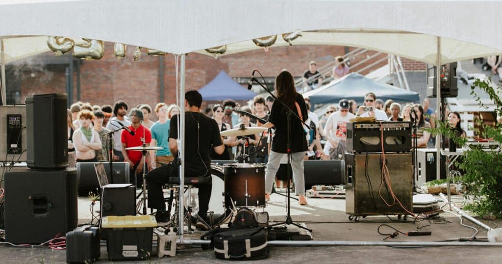 Live outdoor concert performance at a music festival with band playing under a tent, audience enjoying the music, vibrant atmosphere, and sound equipment setup.