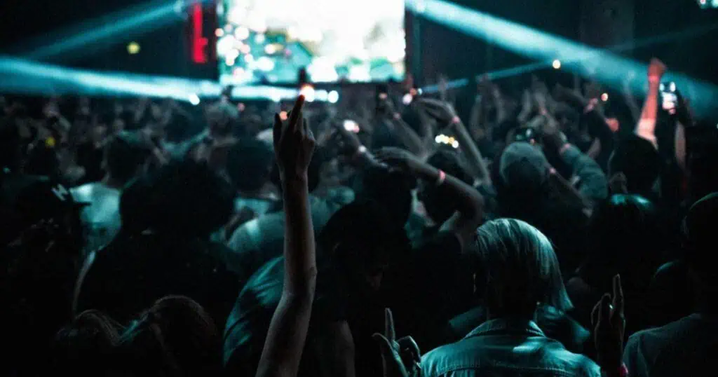 Živahna koncertna publika na glazbenom događaju uživo s podignutim rukama, raznobojnim svjetlima i energičnom atmosferom, koja dočarava bit glazbenih festivala i zabave u noćnom životu.