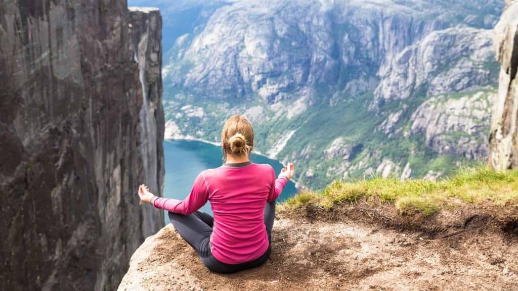 Woman in pink shirt meditating on cliff overlooking scenic mountain landscape