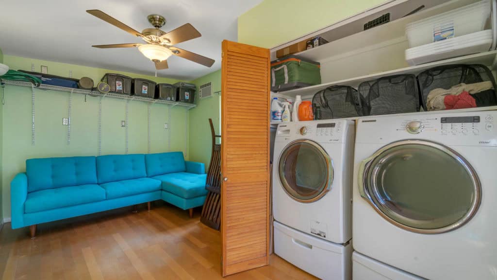 Versatile laundry and storage room at 2212 Myrtle Ave in North Park featuring a high-efficiency front-loading washer and dryer, lime green walls, and a teal sectional sofa under a ceiling fan.