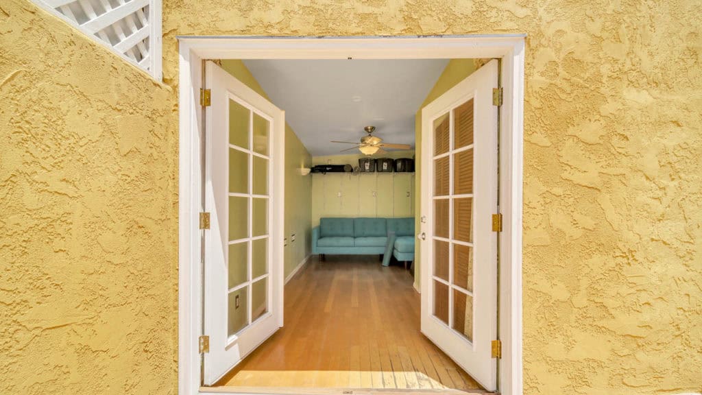 View through open white French doors at 2212 Myrtle Ave in North Park leading into a bright studio or living space with hardwood floors and a teal sectional sofa.