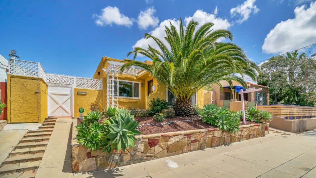 Golden-yellow Spanish Revival home at 2212 Myrtle Ave in North Park featuring a white trellis, decorative stone retaining wall, and lush desert landscaping under a bright blue sky with scattered clouds.