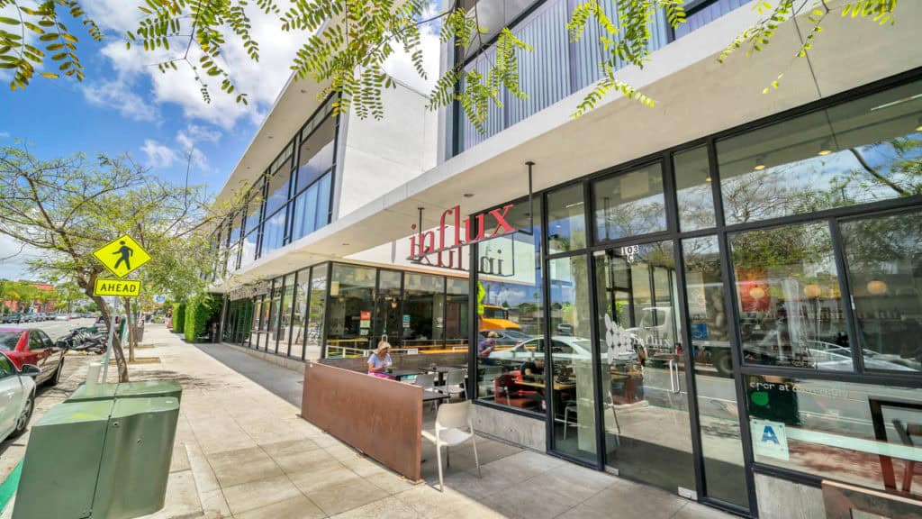 Modern storefront for Influx Cafe in North Park featuring floor-to-ceiling glass windows, a small outdoor seating area with a rusted metal planter, and a wide concrete sidewalk.