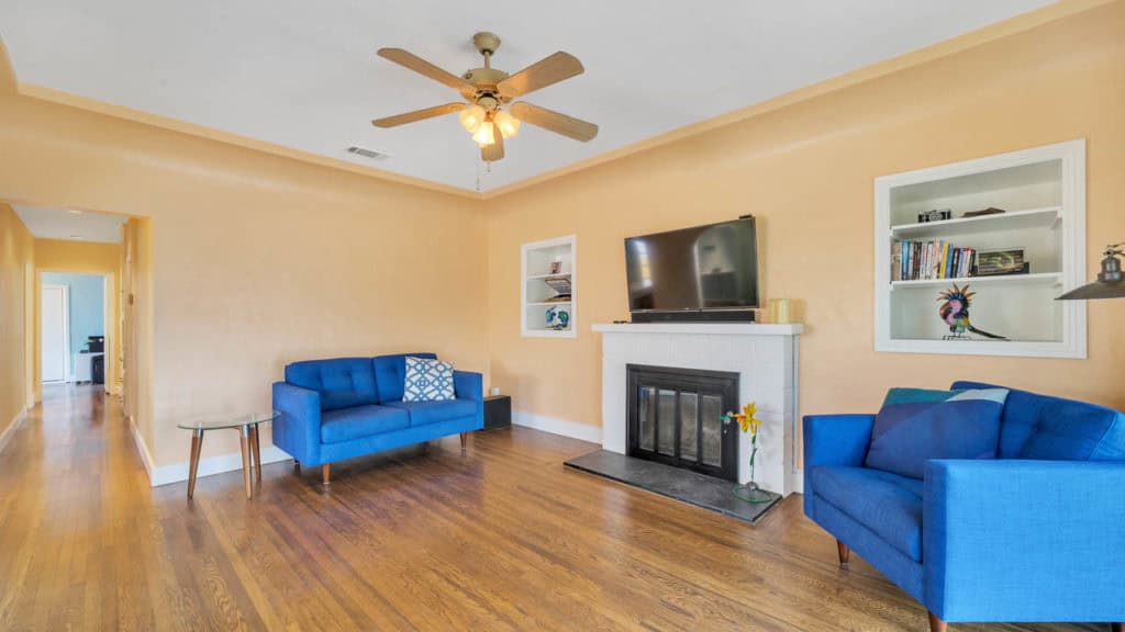 Living room at 2212 Myrtle Ave in North Park featuring warm peach-colored walls, hardwood floors, a white fireplace with a mounted TV, and vibrant blue mid-century modern seating.
