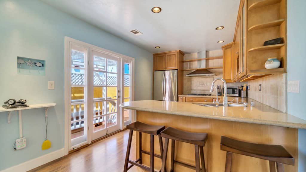 Modern kitchen at 2212 Myrtle Ave in North Park featuring light blue walls, wood cabinetry, stainless steel appliances, and a breakfast bar with wood stools next to French doors leading to the patio.