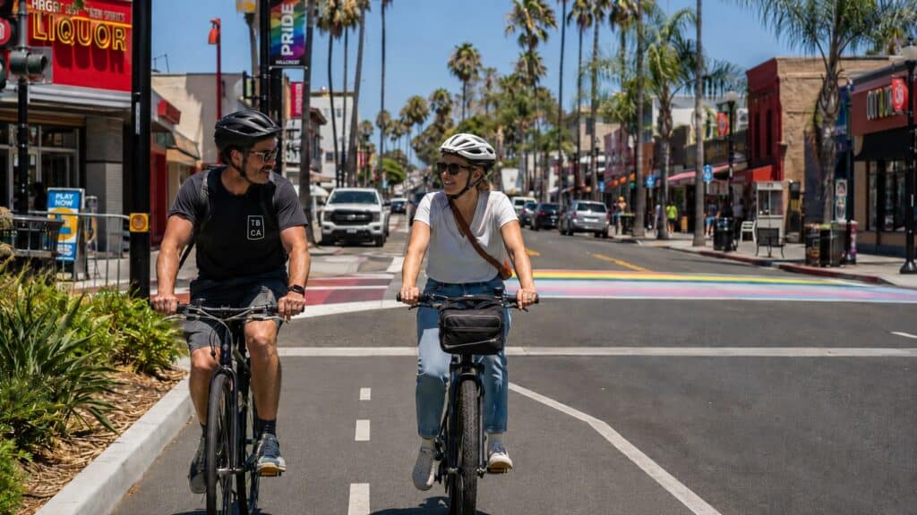 Man and woman in their 30s ride commuter bikes side by side in a protected bike lane on University Avenue in Hillcrest, San Diego, with palm trees, colorful storefronts, and a rainbow Pride crosswalk in the background