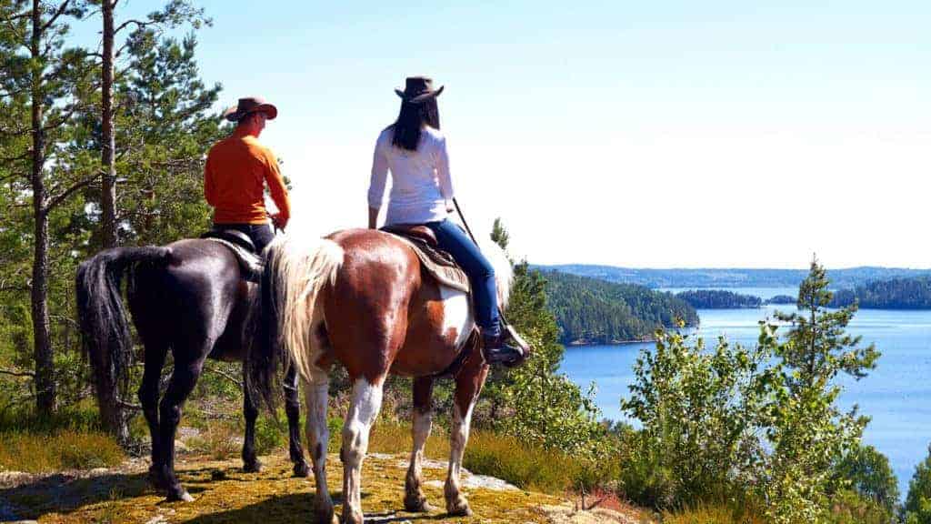 Två personer som sitter på var sin häst står på ett berg. I bakgrunden syns en vacker sjö.
