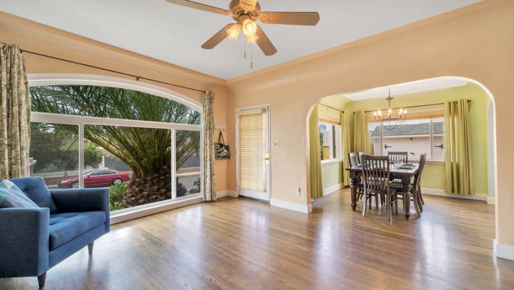 Bright living and dining area at 2212 Myrtle Ave in North Park featuring a large arched picture window, hardwood floors, and a wide interior archway leading to a formal dining room with a classic wood table and chandelier.