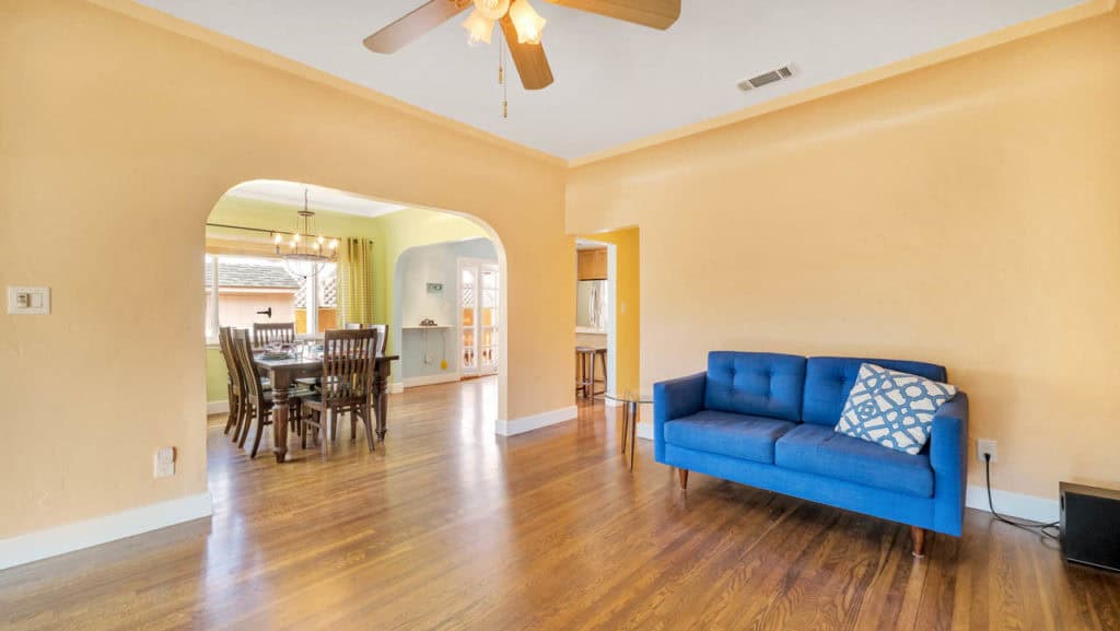Open-concept living space at 2212 Myrtle Ave in North Park with warm peach-colored walls and hardwood floors, showing a blue mid-century modern sofa and an arched doorway leading to the dining room.