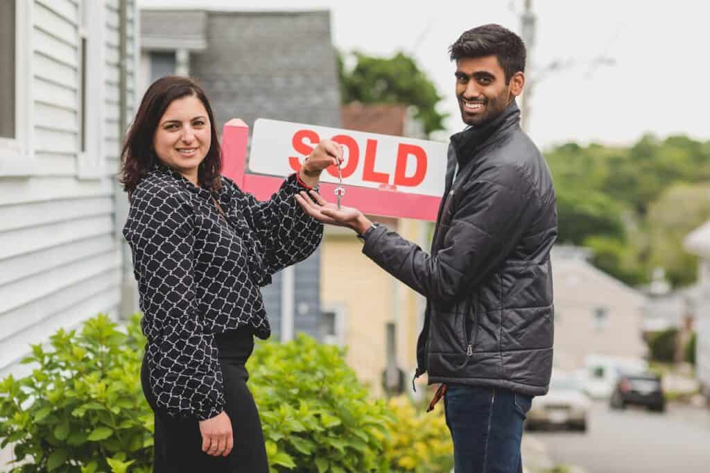 A Realtor Giving a Client a Key to the House with a Sold Sign on the Background