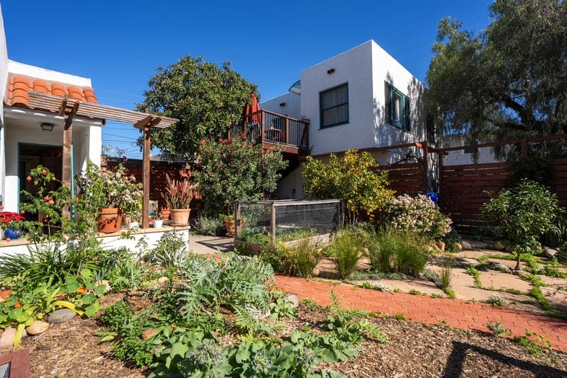 Private backyard at 3237-39 Dale St in North Park, featuring a detached two-story casita with a loquat tree deck, a raised garden bed, and a red brick walkway surrounded by mature Mediterranean landscaping.