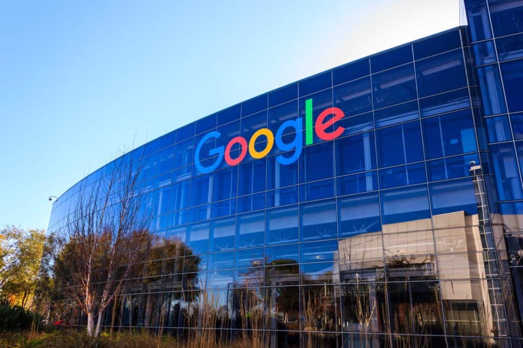 Edifício curvo de vidro azul da Google com o logotipo colorido, refletindo o céu e árvores em um dia ensolarado.