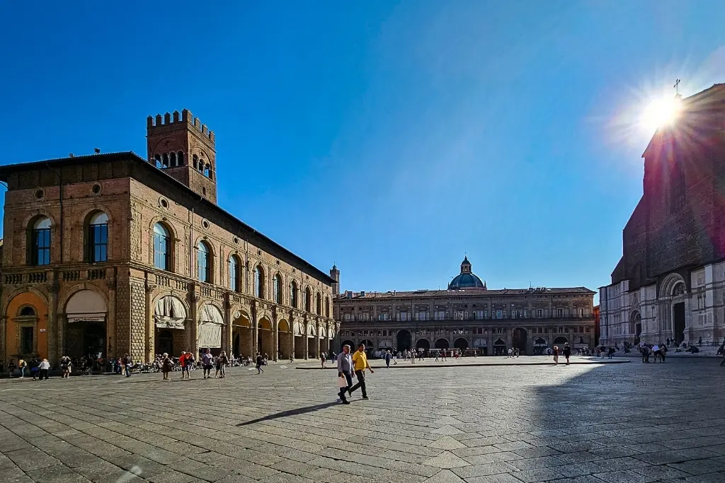 Wide-angle photo of Piazza Maggiore in Bologna with Palazzo dei Banchi, Basilica di San Petronio, and sun flare—perfect for capturing the city's main square and daily rhythm.