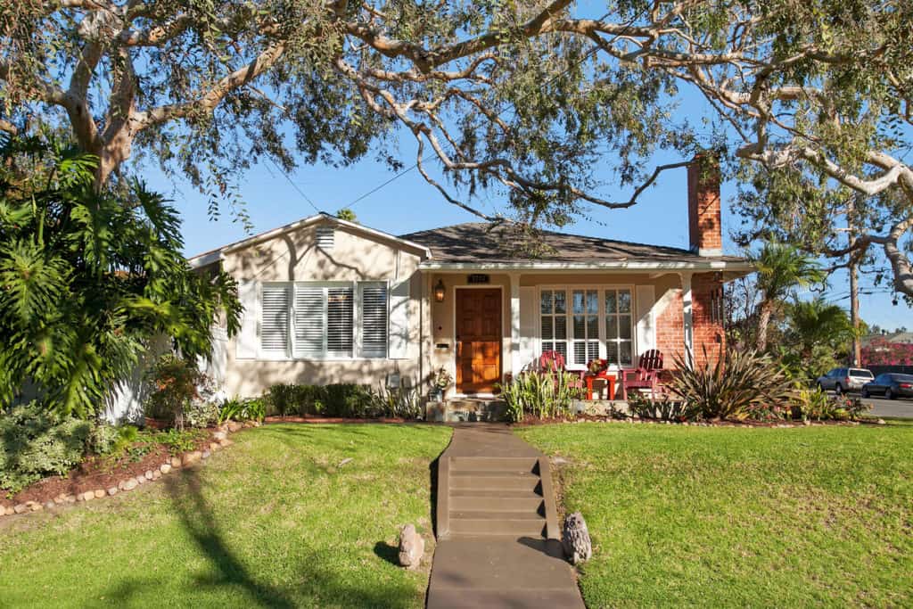 Beige Craftsman-style home in North Park with a brick chimney, wooden front door, and a green lawn shaded by large trees.