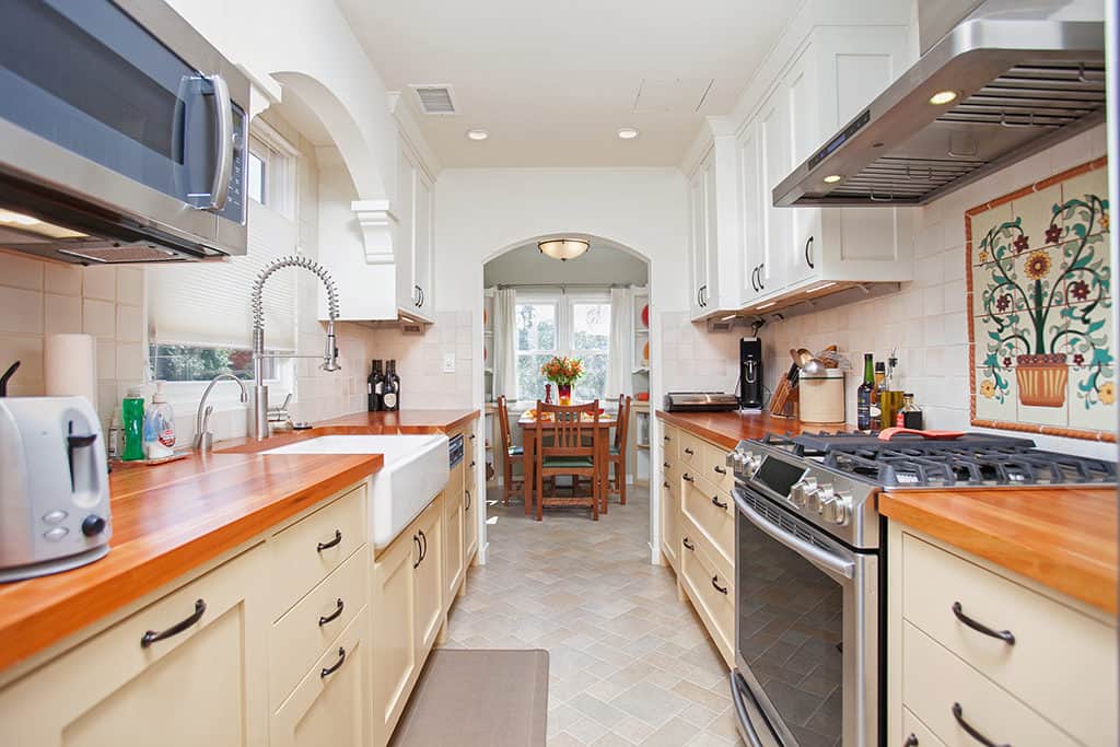 Modern galley kitchen at 3317-3319 Boundary Street in North Park featuring cream-colored cabinetry, warm wood countertops, and a large white farmhouse sink, with an arched doorway leading to the dining room.