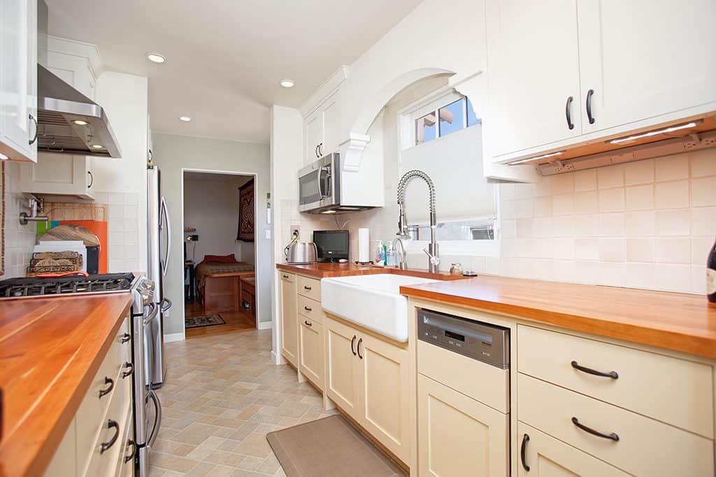 Modern galley kitchen at 3317-3319 Boundary Street in North Park featuring cream-colored cabinetry, warm wood countertops, a large white farmhouse sink, and stainless steel appliances.