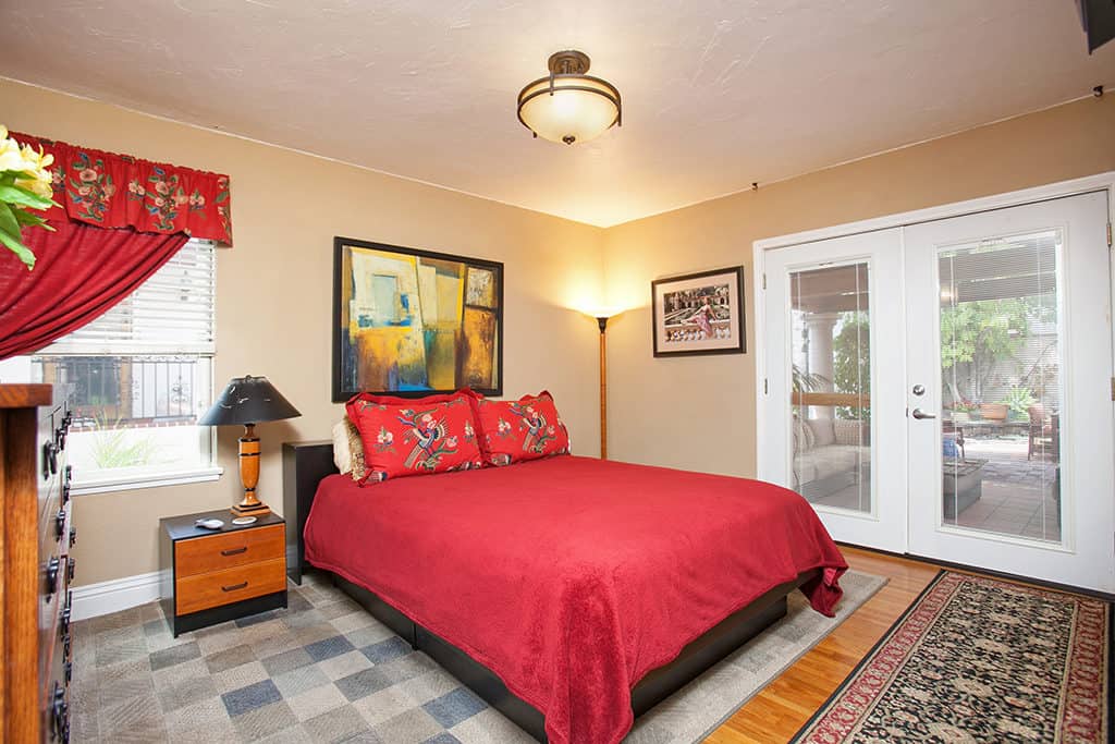 Bedroom at 3317-3319 Boundary Street in North Park featuring a vibrant red bedspread and pillows, neutral beige walls with abstract artwork, and glass French doors leading to a private outdoor patio.