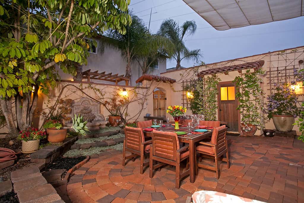 Outdoor dining courtyard at 3317-3319 Boundary Street in North Park featuring a wood table set, circular brick paver patio, a stone tiered fountain, and lush climbing vines against a Mediterranean-style wall.