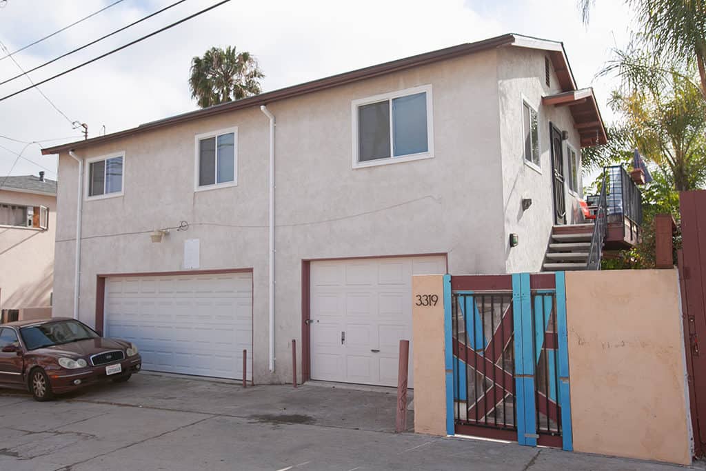 Two-story residential building at 3317-3319 Boundary Street in North Park featuring two white garage doors, an exterior staircase leading to a second-floor unit, and a unique blue and red patterned gate.