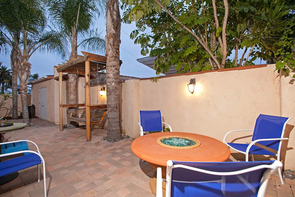 Private paved patio at 3317-3319 Boundary Street in North Park featuring a circular fire pit with blue glass, vibrant blue patio chairs, a wooden porch swing under a pergola, and tropical palm trees.
