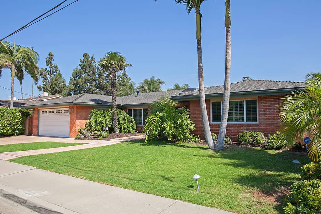 Single-story brick ranch home at 6253 Cypress Point Rd in Del Cerro featuring a manicured green lawn, lush tropical landscaping with palm trees, and a white two-car garage.