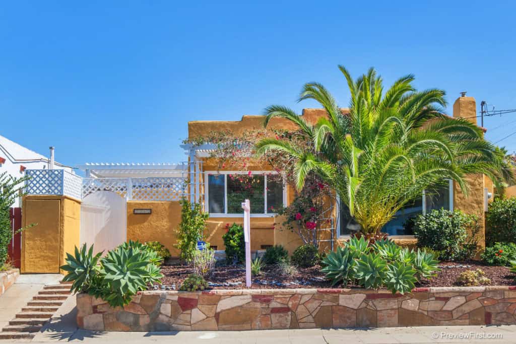 Spanish Revival-style bungalow with golden-yellow stucco at 2212 Myrtle Ave in North Park, featuring a stone retaining wall, desert landscaping, and a lush palm tree under a clear blue sky.
