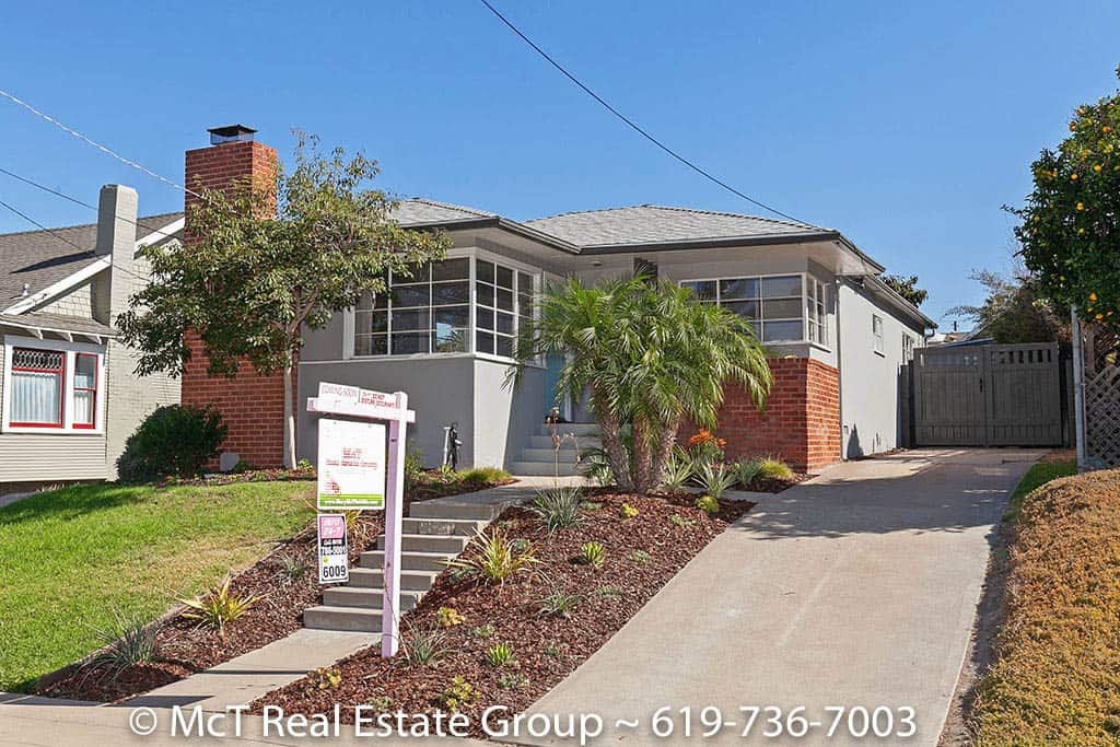 Exterior view of a contemporary grey home at 1627 Granada Avenue in South Park featuring a prominent red brick chimney, wrap-around corner windows, a tiered garden bed with succulents, and a paved driveway under a clear sky.