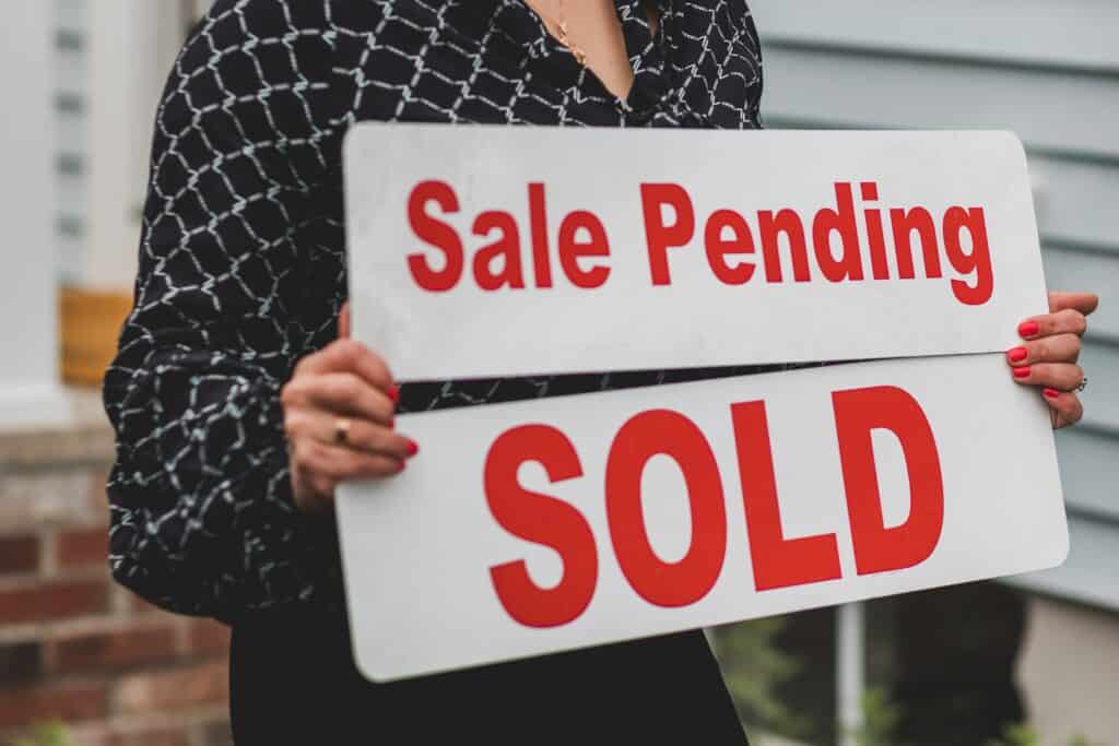 A person with red nail polish holding two white real estate signs that read "Sale Pending" and "SOLD" in red text in front of a house.