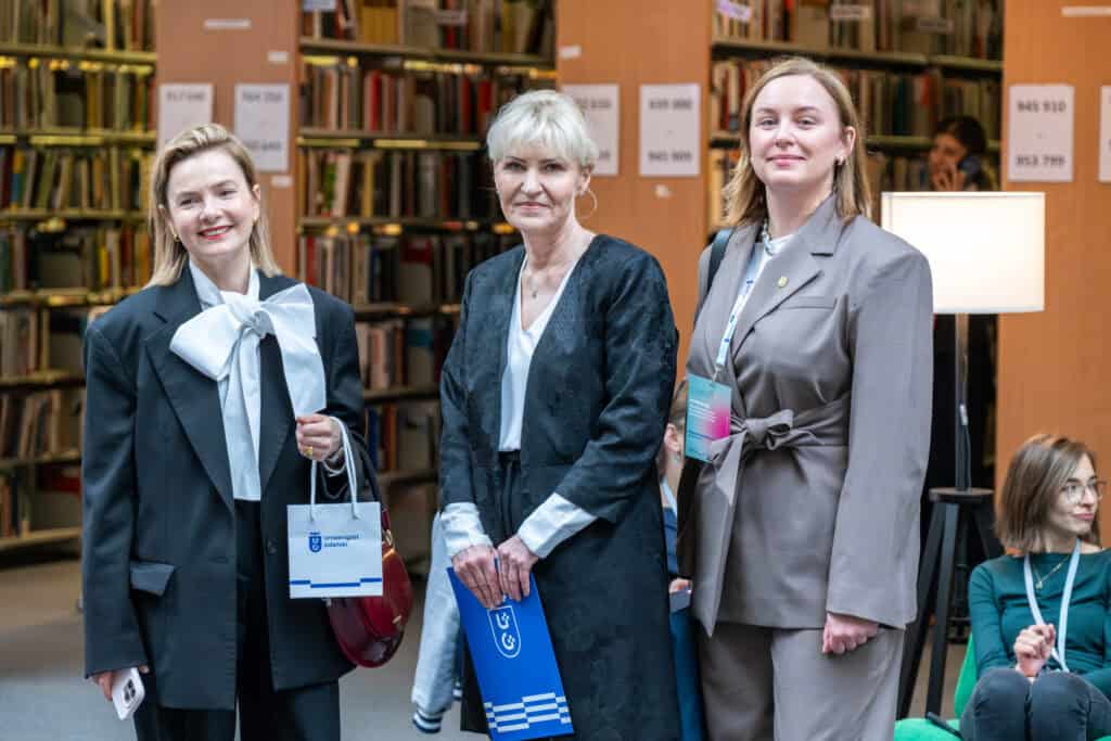 trzy kobiety pozujące do zdjęcia na stojąco na tle półek z książkami