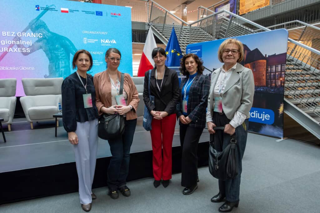 Grupa pięciu kobiet na konferencji EURAXESS, stojąca na scenie z flagami Polski i UE w tle, w nowoczesnym centrum konferencyjnym.