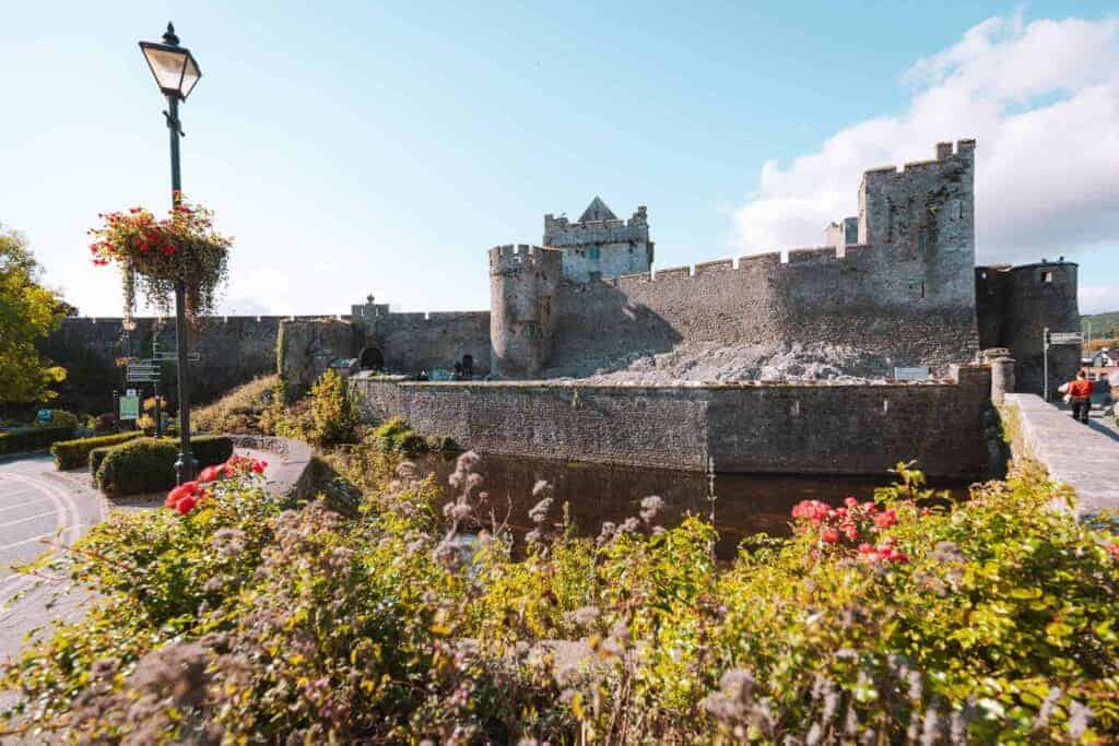 Exterior of a medieval stone castle, surrounded by a moat, under a bright blue sky. The castle, with its high walls and round towers, appears to be well-preserved, and a streetlamp with hanging flower baskets is seen in the foreground, along with vibrant greenery and red flowers.
