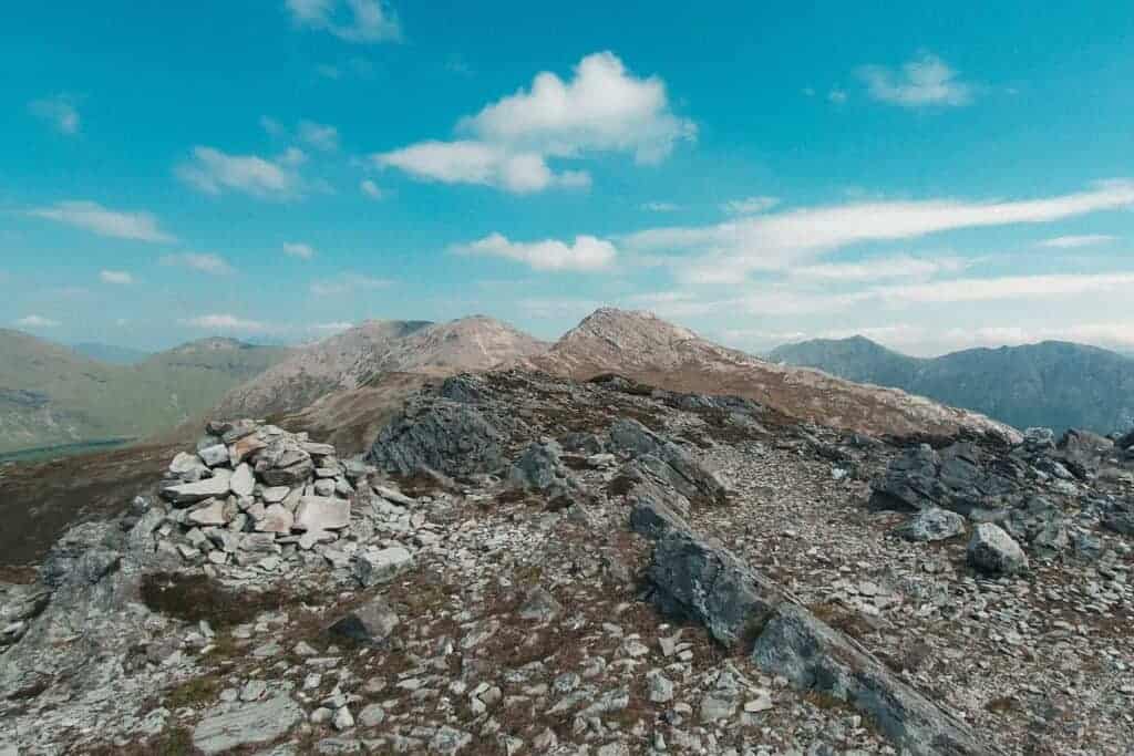 A pile of stones marking the summit of Benlettery, with rugged, rocky mountain peaks in the background and a bright blue sky overhead.