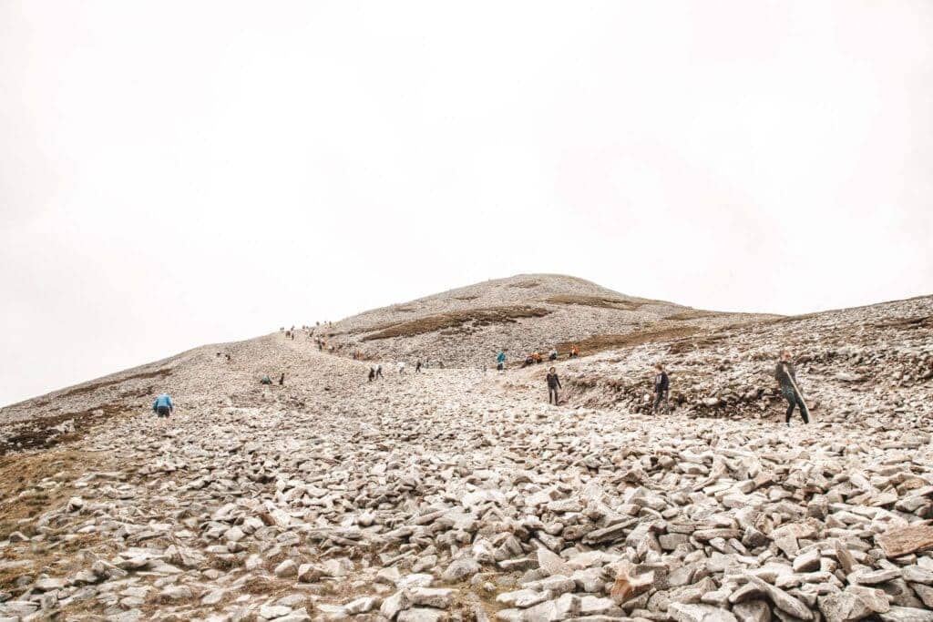 The summit of Croagh Patrick with hikers scattered across a rocky terrain, climbing towards the peak under an overcast sky.