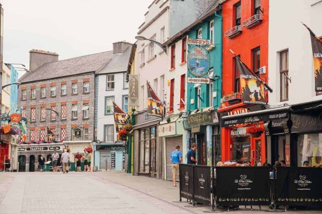 Colorful storefronts and flags adorn the lively streets of Galway, with people walking and chatting under a partly cloudy sky.